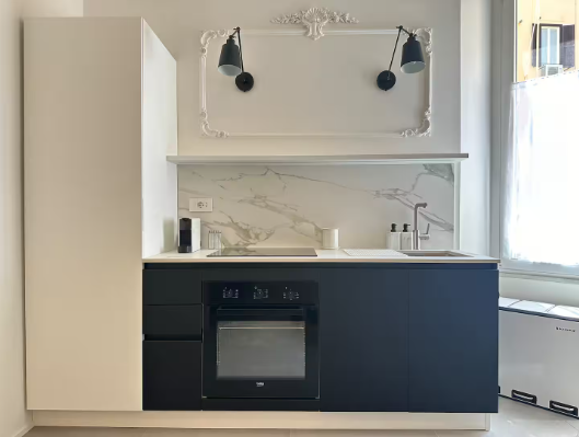 Piccola cucina moderna con mobili bianchi e neri, paraschizzi in marmo e due applique nere.