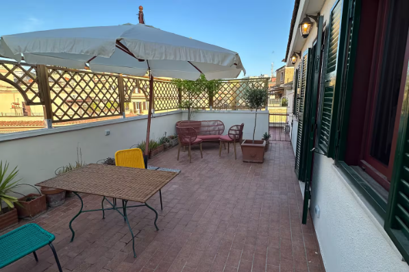 Patio sul tetto con tavolo, sedie, piante e ombrellone, caratterizzato da pavimento in piastrelle rosse e vista sugli edifici.