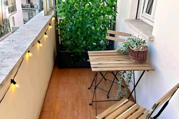 Un piccolo balcone con un tavolo di legno, due sedie e una pianta in vaso, con luci a filo.