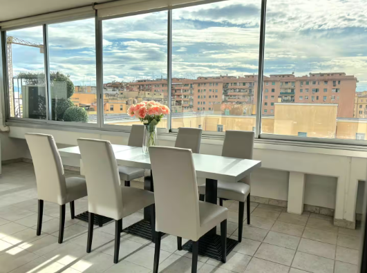 Sala da pranzo con tavolo bianco, sei sedie grigie e grandi finestre con vista sulla città.