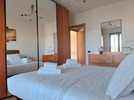 Camera da letto con letto, cassettiera, specchio e armadio. Sul letto sono presenti lenzuola e asciugamani bianchi.