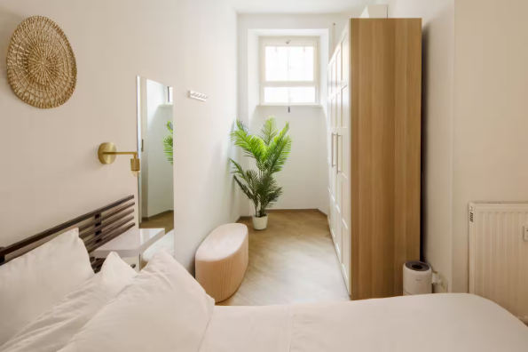 Camera da letto con letto, ampio armadio, specchio e una piccola panca. Finestra e pianta a vista.