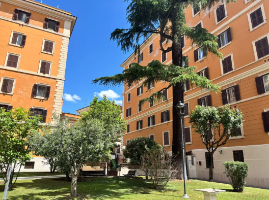 Edifici con muri arancioni e persiane marroni circondano un cortile verde con alberi e un cielo azzurro.
