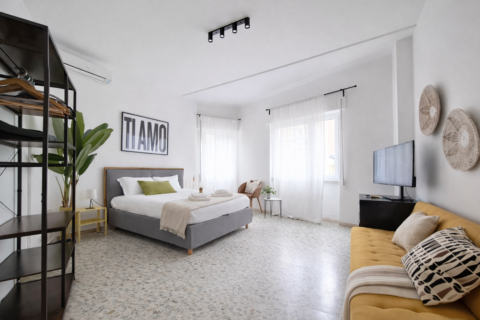 Monolocale luminoso e moderno con letto, divano, TV e libreria a vista. Pareti bianche e pavimenti in terrazzo.