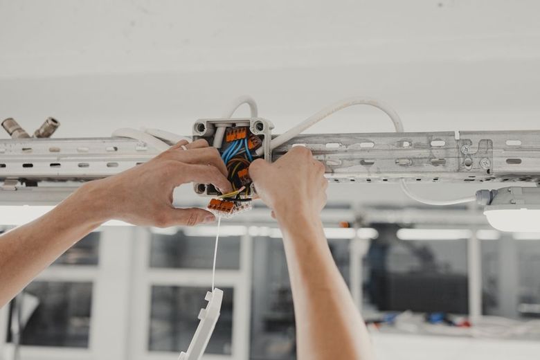 Hands wiring electrical box on a ceiling; wires, metal rail, and a bright interior setting.