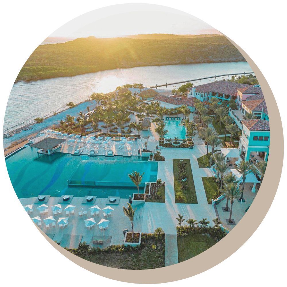 Aerial view of Sandals resort Curacao with turquoise pools, white umbrellas, and buildings, near a body of water, at sunset.