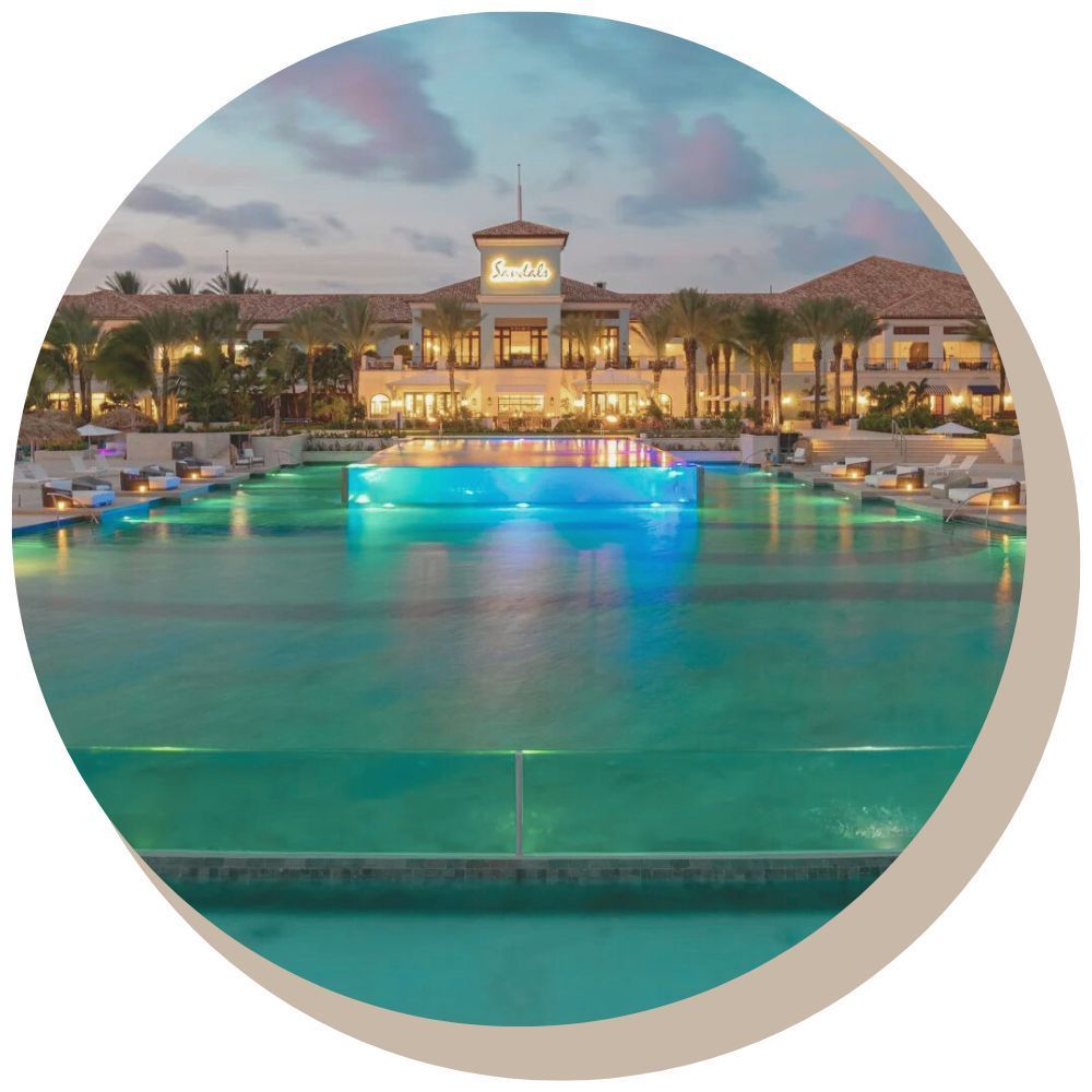 Resort pool at dusk with illuminated water features and a building in the background.