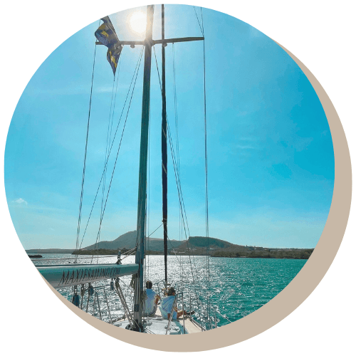 Sailboat on turquoise water with sun, mast, two people, and land in distance.