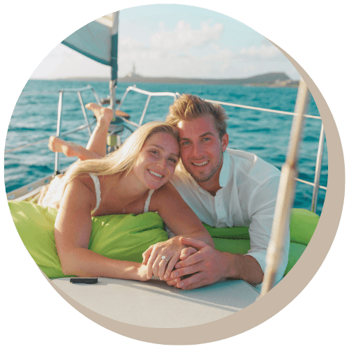Couple on a sailboat, smiling. They hold hands, with ocean and distant land in the background.