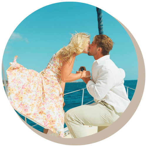 Couple kissing on a sailboat, the man is kneeling, proposing with a ring, against a blue sky.