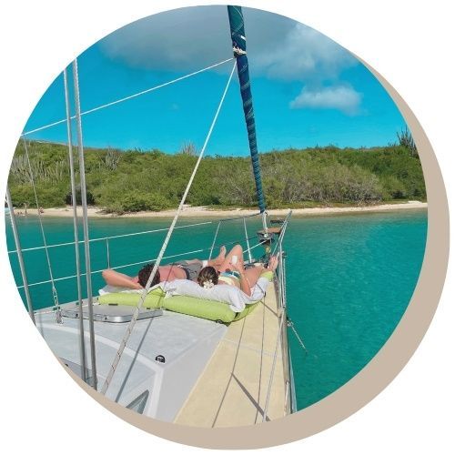 Two people enjoying a private Maxie Sailing Curacao tour - relaxing on a boat's deck, in turquoise waters of Fuik Bay and shoreline in the background.