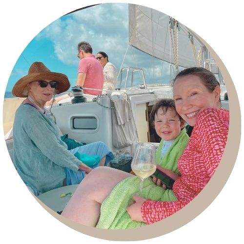 Happy family on sailboat Galaxie on Caribbean Sea, woman in red top smiles, child wrapped in towel, mother and grandmother visible.