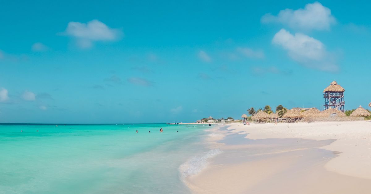 Turquoise ocean at Klein Curacao white sand beach under a clear blue sky. A tower and huts visible.