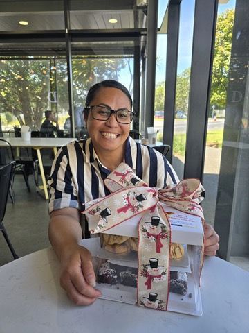 Woman smiles, holding a gift box of treats in cafe setting. Snowman ribbon, striped shirt, glasses.