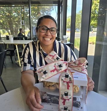 Woman with glasses smiling, holding a gift box tied with a snowman ribbon, sitting in a cafe.