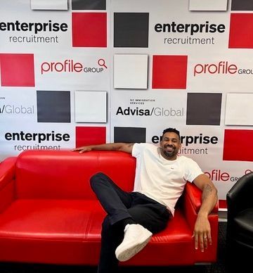 Man sitting on a red sofa, smiling, in front of a wall with recruitment company logos.