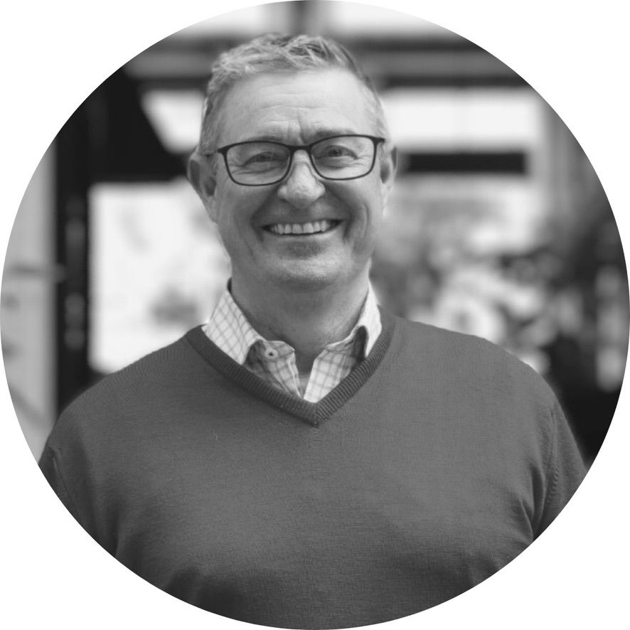 Man with glasses smiles, wearing a sweater over a collared shirt.