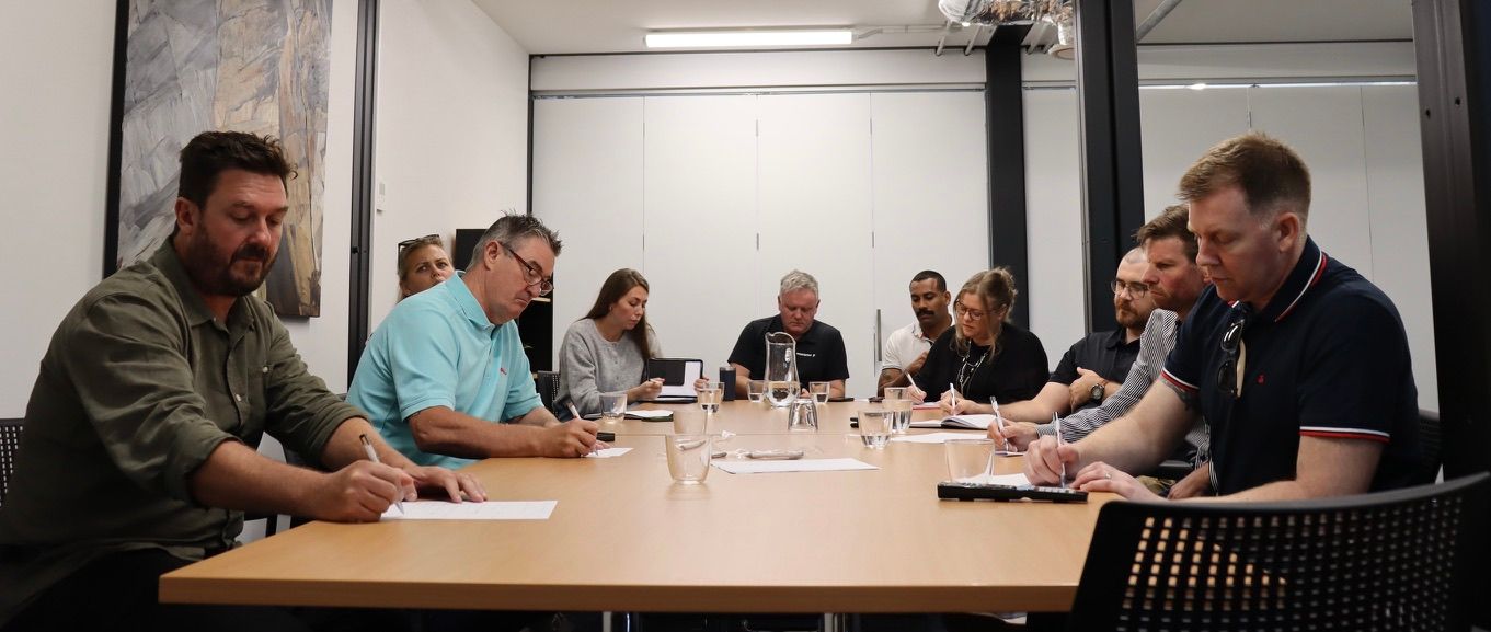 People seated around a conference table writing.  They are in a brightly lit room.