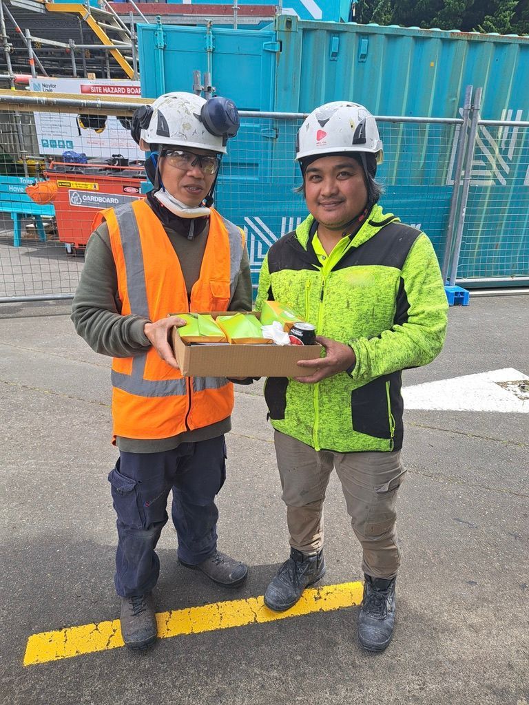Two construction workers holding a box of food outdoors near a building site. One wears orange vest, other a green jacket.