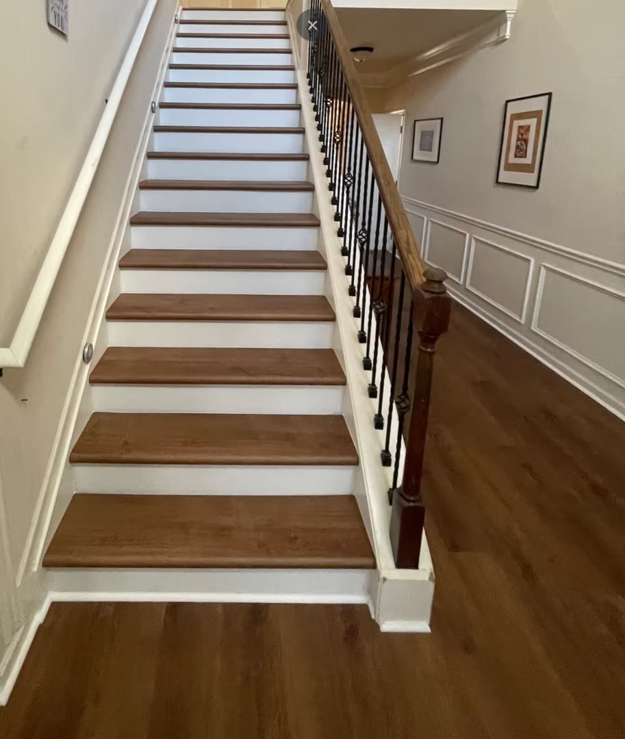 Staircase with wood steps and white trim. Wooden handrail with black spindles. Hallway with wood flooring.