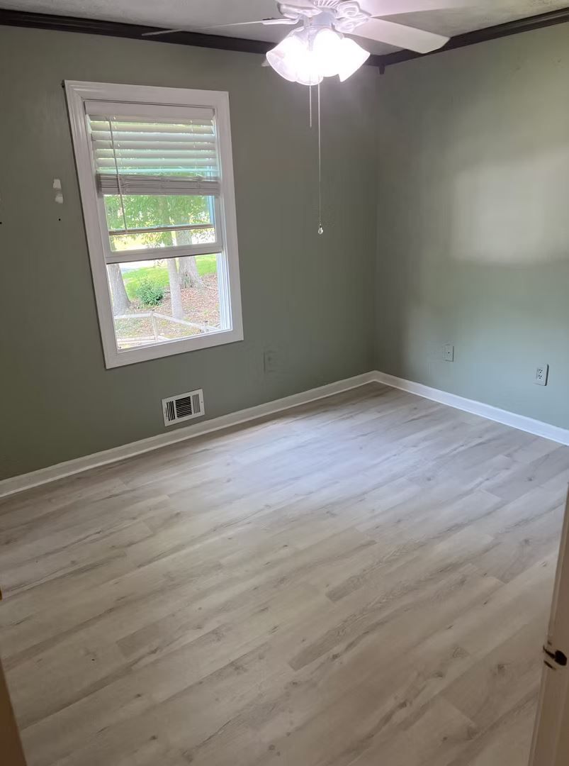 Empty bedroom with light wood-look flooring and sage green walls, a window with blinds, and a ceiling fan.