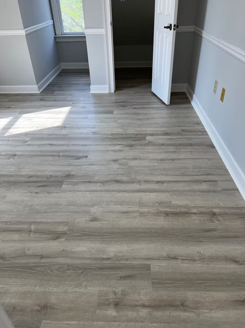 Room with gray wood-look flooring, white trim, and an open door leading to another space.