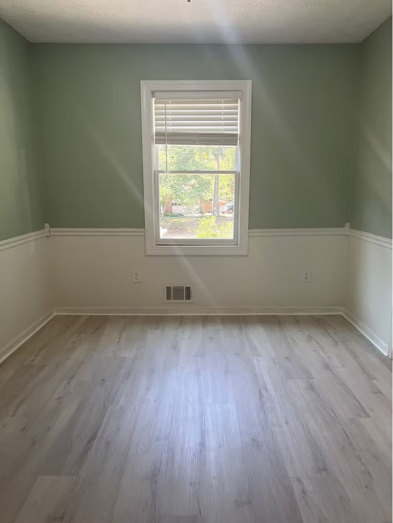 Empty room with light wood-look flooring, white wainscoting, and pale green walls. A window is centered on the wall.