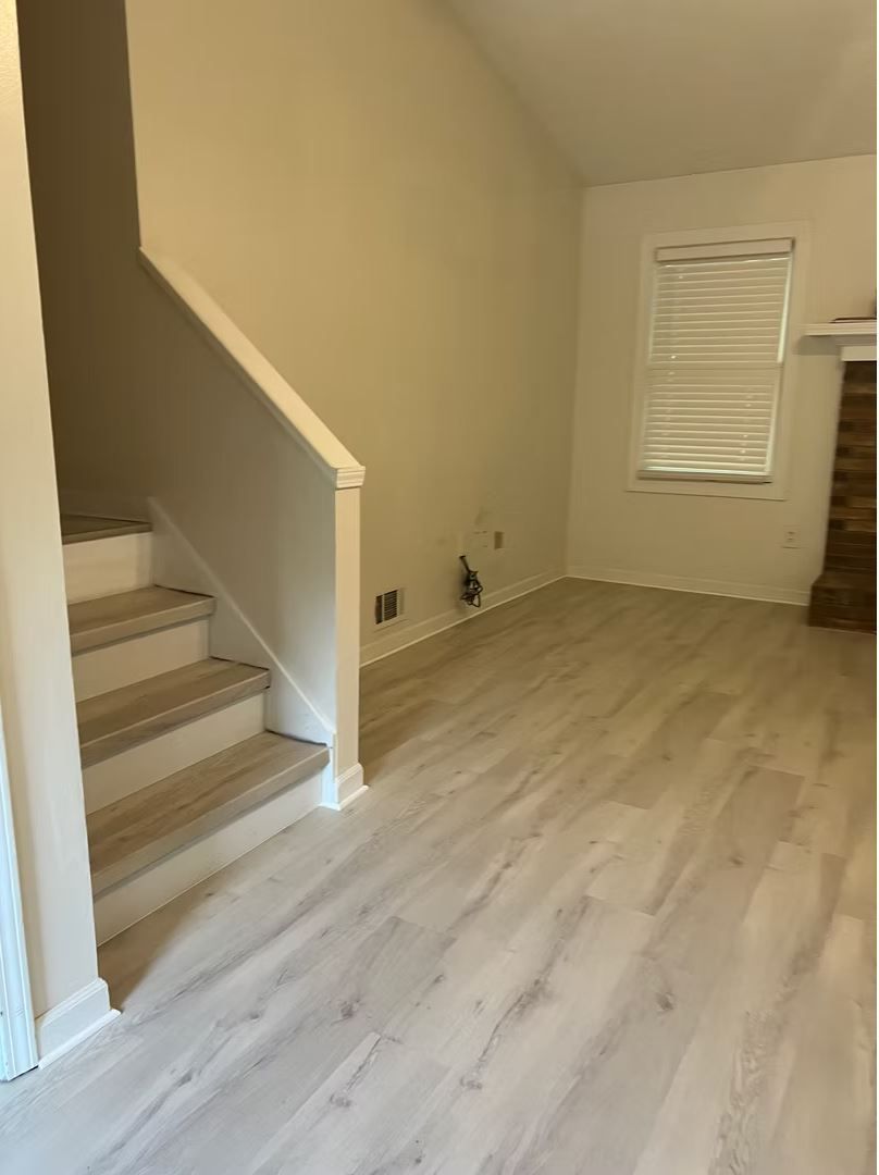 A bright, empty room with stairs and a window. Light wood-look flooring and beige walls.