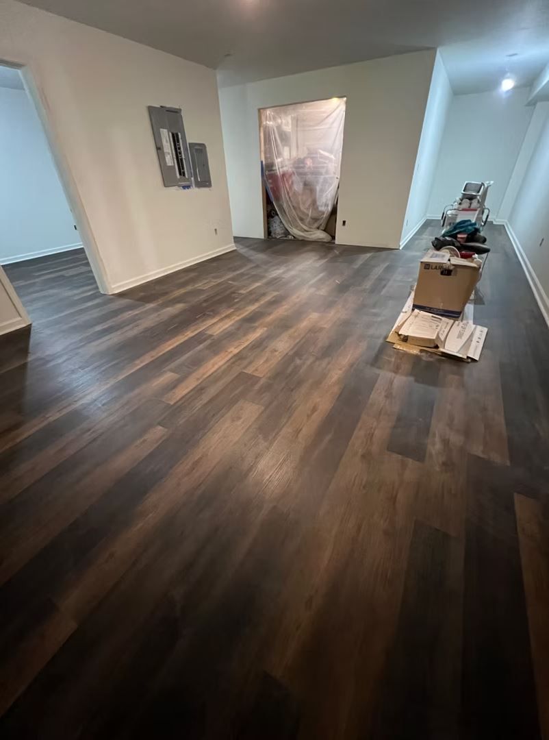 Dark wood-look flooring in a room with white walls; doorway covered with plastic, boxes and tools present.