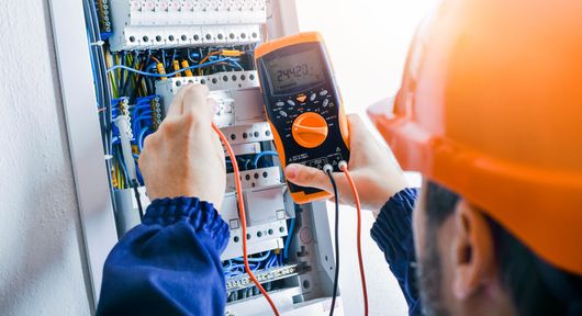 An electrician is working on an electrical panel with a multimeter.