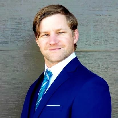 Man in blue suit, white shirt, and turquoise tie smiles in front of a neutral-toned wall.