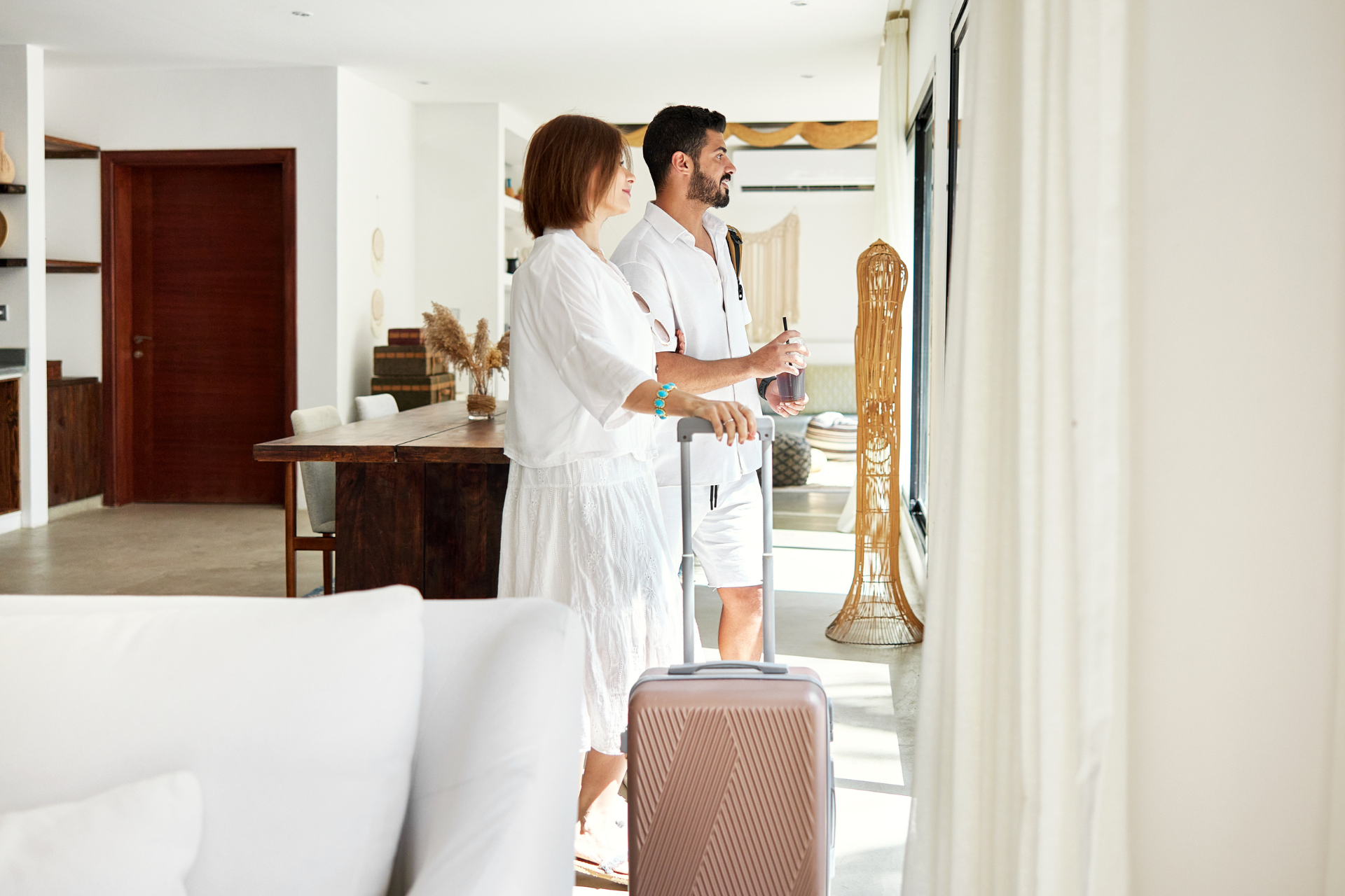 Couple in white outfits looking out a window near their suitcase in a bright room.