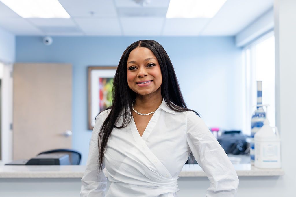 Woman smiles in a white shirt behind a reception desk, light blue wall, and artwork.