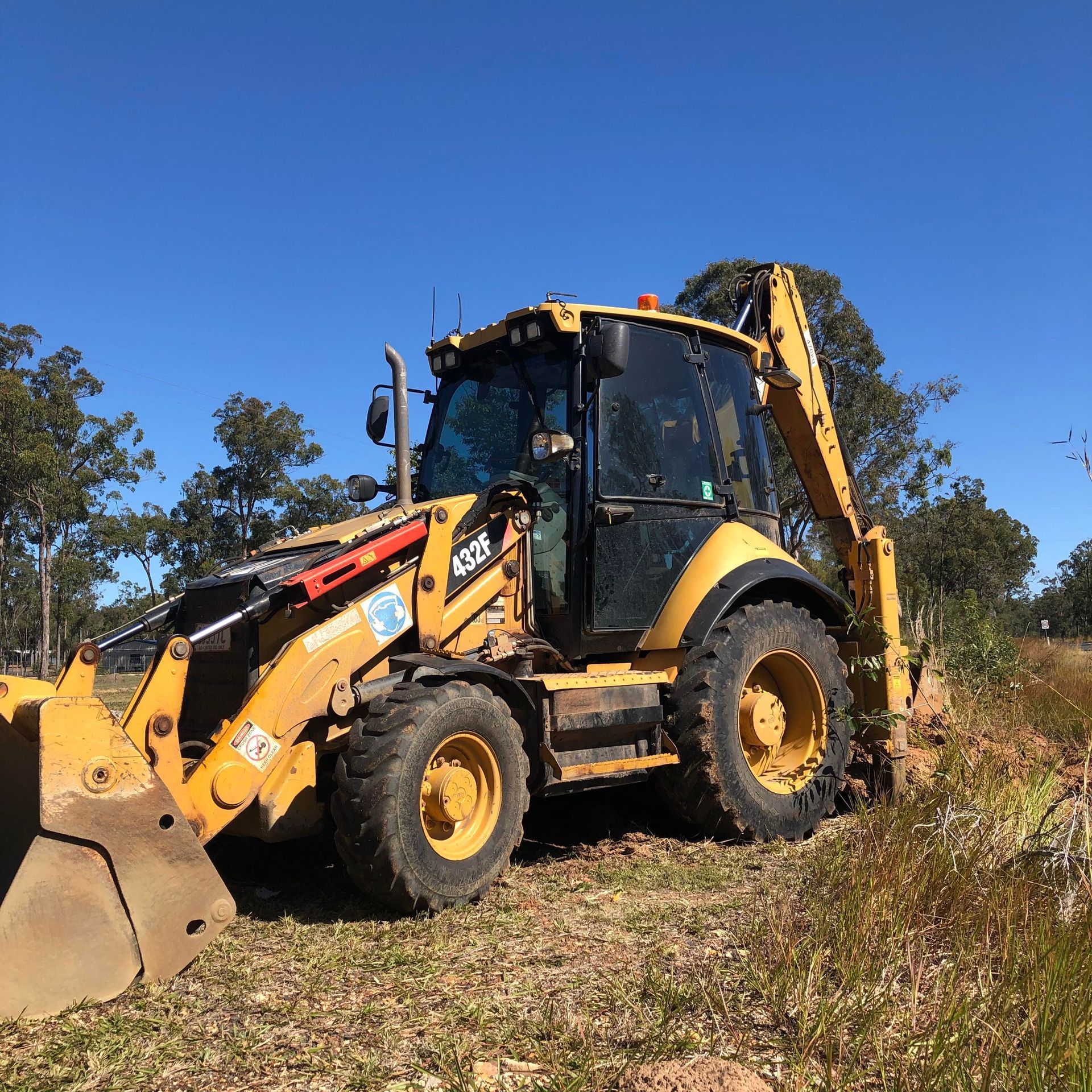 Yellow backhoe excavator on a dirt path against a blue sky, digging near trees.