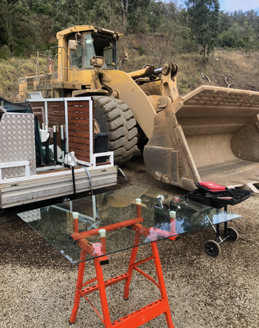A yellow loader next to a trailer with a glass table on a sawhorse outdoors.