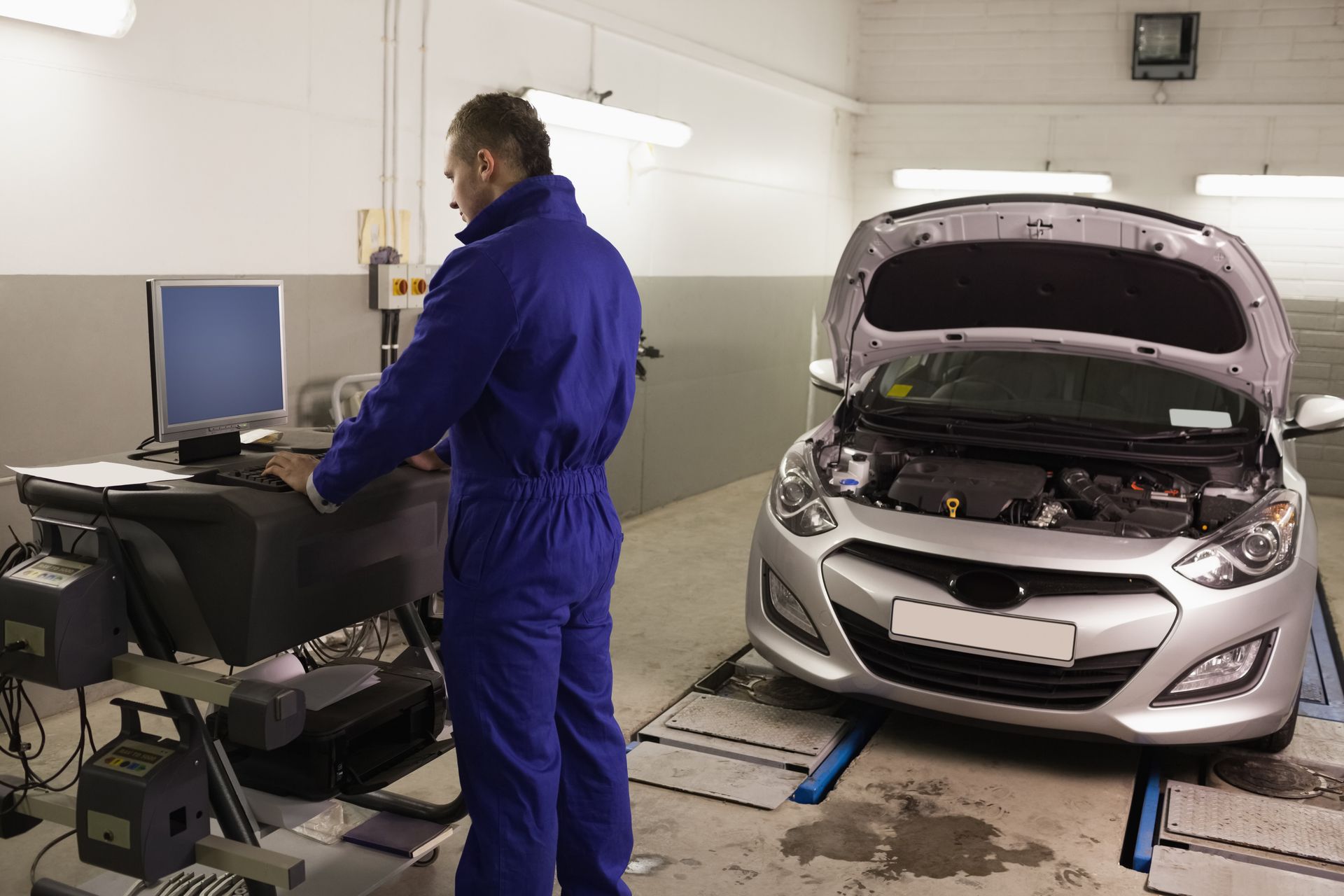 Mechanic in blue jumpsuit using a computer, inspecting a silver car with its hood open in a garage.