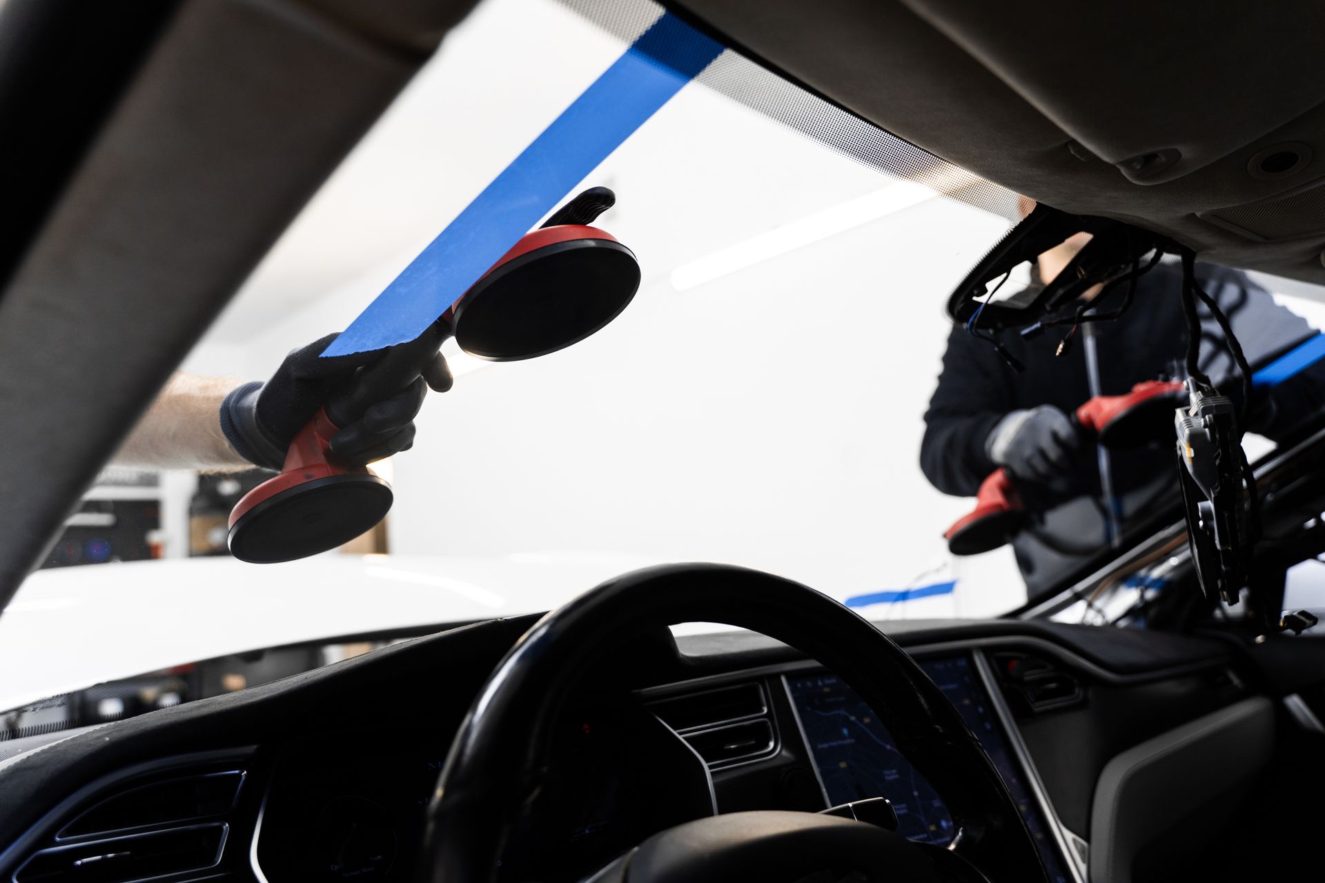 Person replacing a car windshield. A black and red suction cup tool is attached to the glass. The steering wheel is visible.