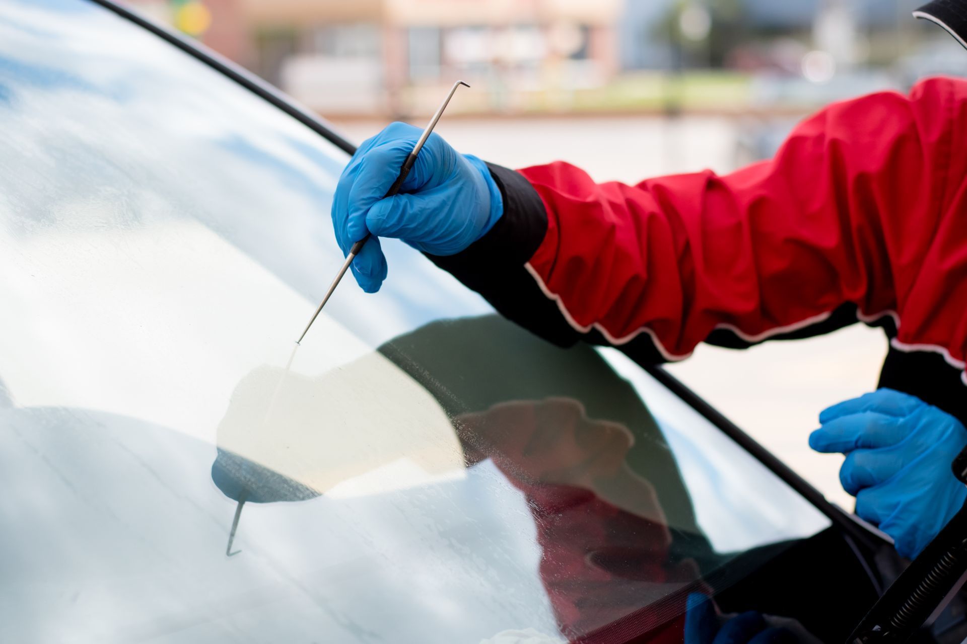 Person in red jacket and blue gloves inspecting a car windshield with a tool outdoors.