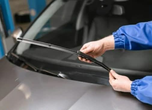 Person in blue uniform replacing a windshield wiper on a gray car.