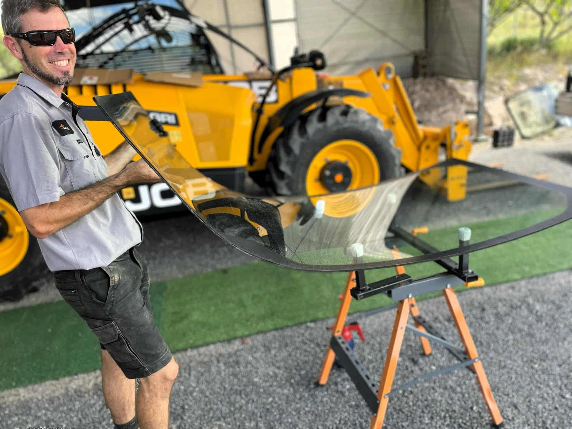 Man holding curved glass, smiling, with yellow JCB tractor in background.