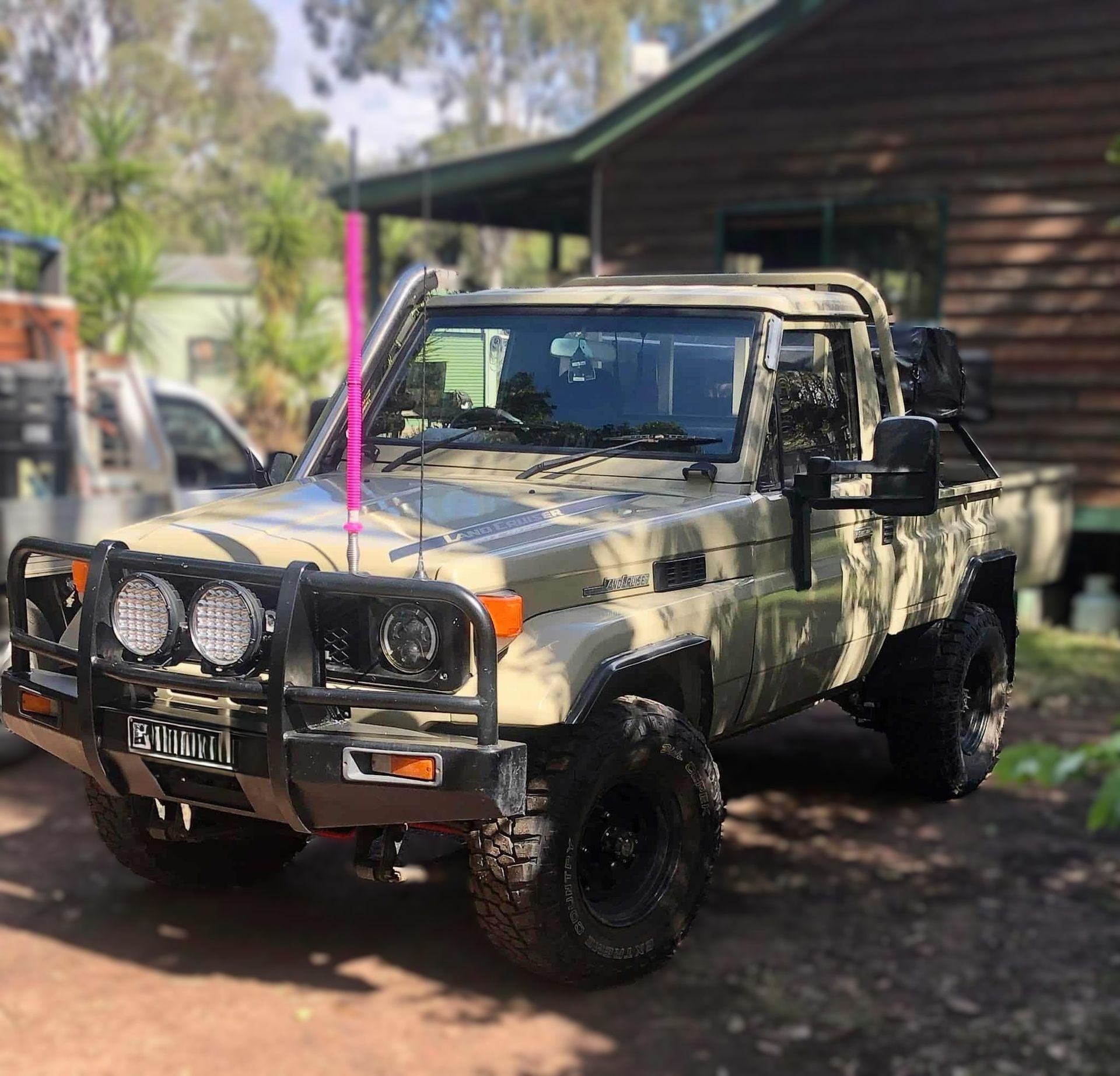 Tan off-road truck parked in front of a wooden cabin. It has a bull bar, spotlights, and large tires.