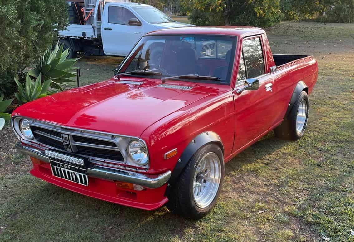 Red, modified classic Datsun pickup truck with wide tires, on grass.