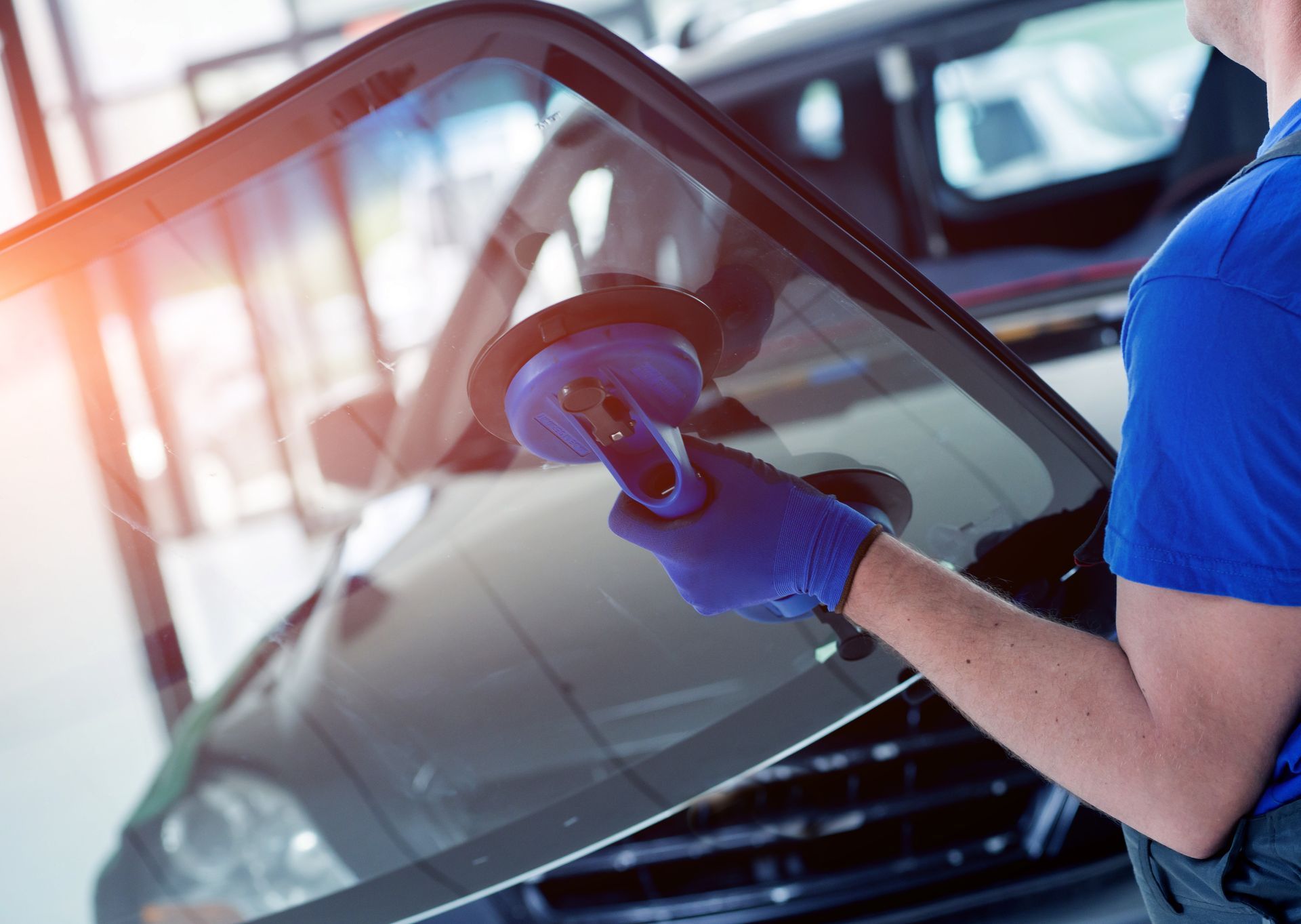 Mechanic using a suction cup to remove a car windshield, blue gloves, sunny workshop.