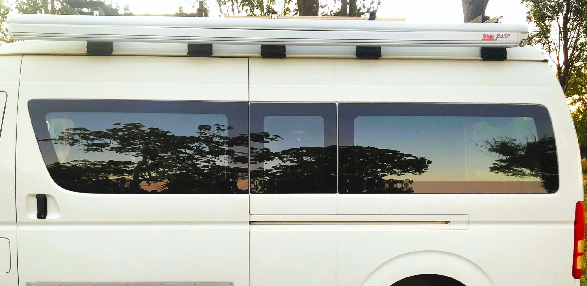 White van with tinted windows reflecting trees, a rolled-up awning on top.