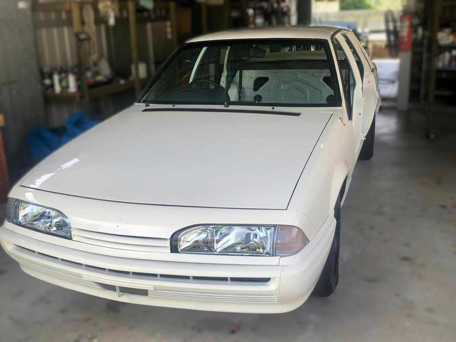 White car, likely a rally build, in a garage. Shows front view with custom lights and a roll cage visible.