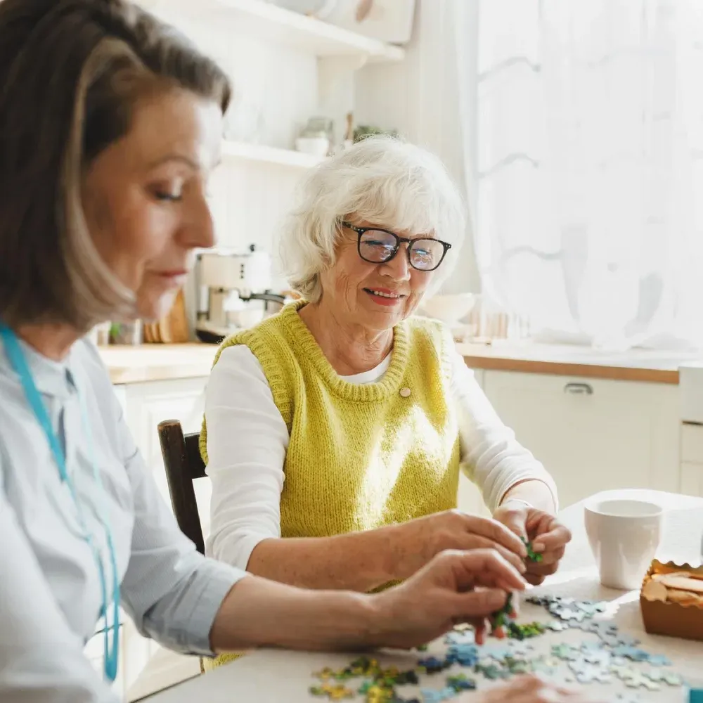 Woman assisting another woman with a puzzle in a bright kitchen.