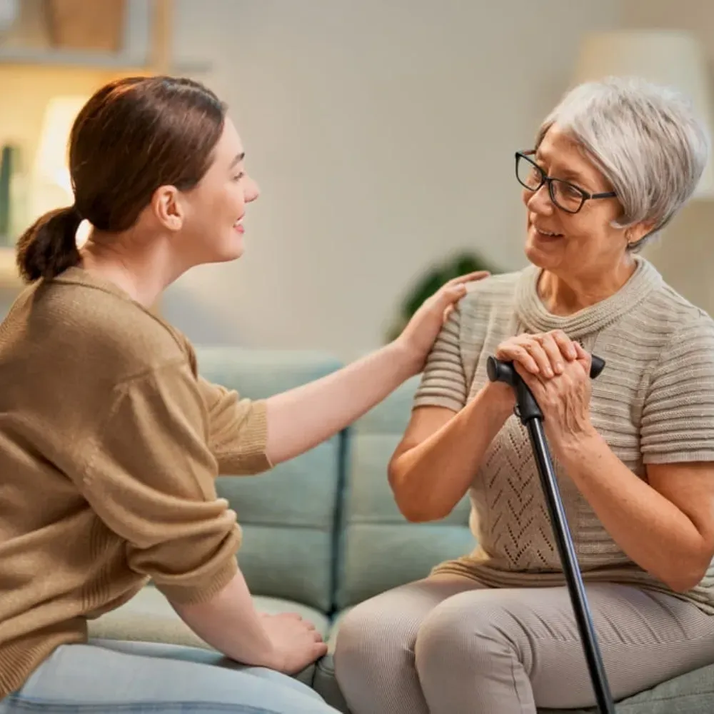 Woman with ponytail comforts another woman seated with a cane, both smiling on a sofa.