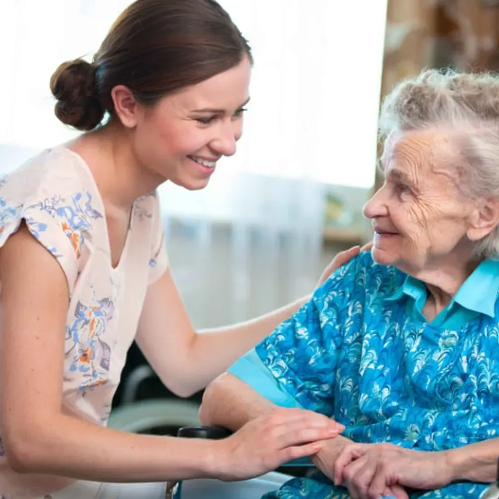 Woman smiles, comforting an elderly person in a wheelchair; both hold hands; indoor setting.