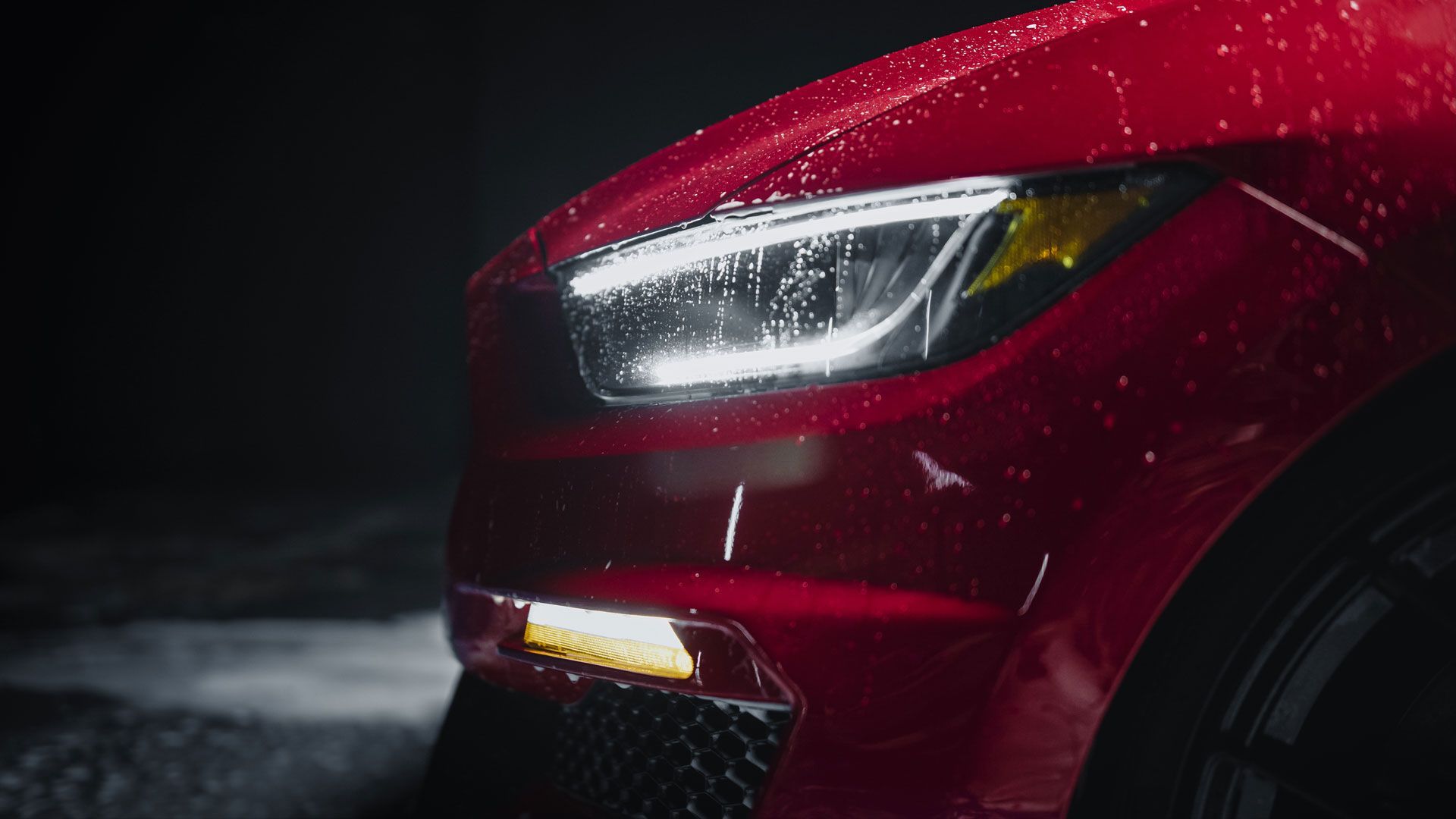 Red sports car headlight illuminated with water droplets, in a dark setting.