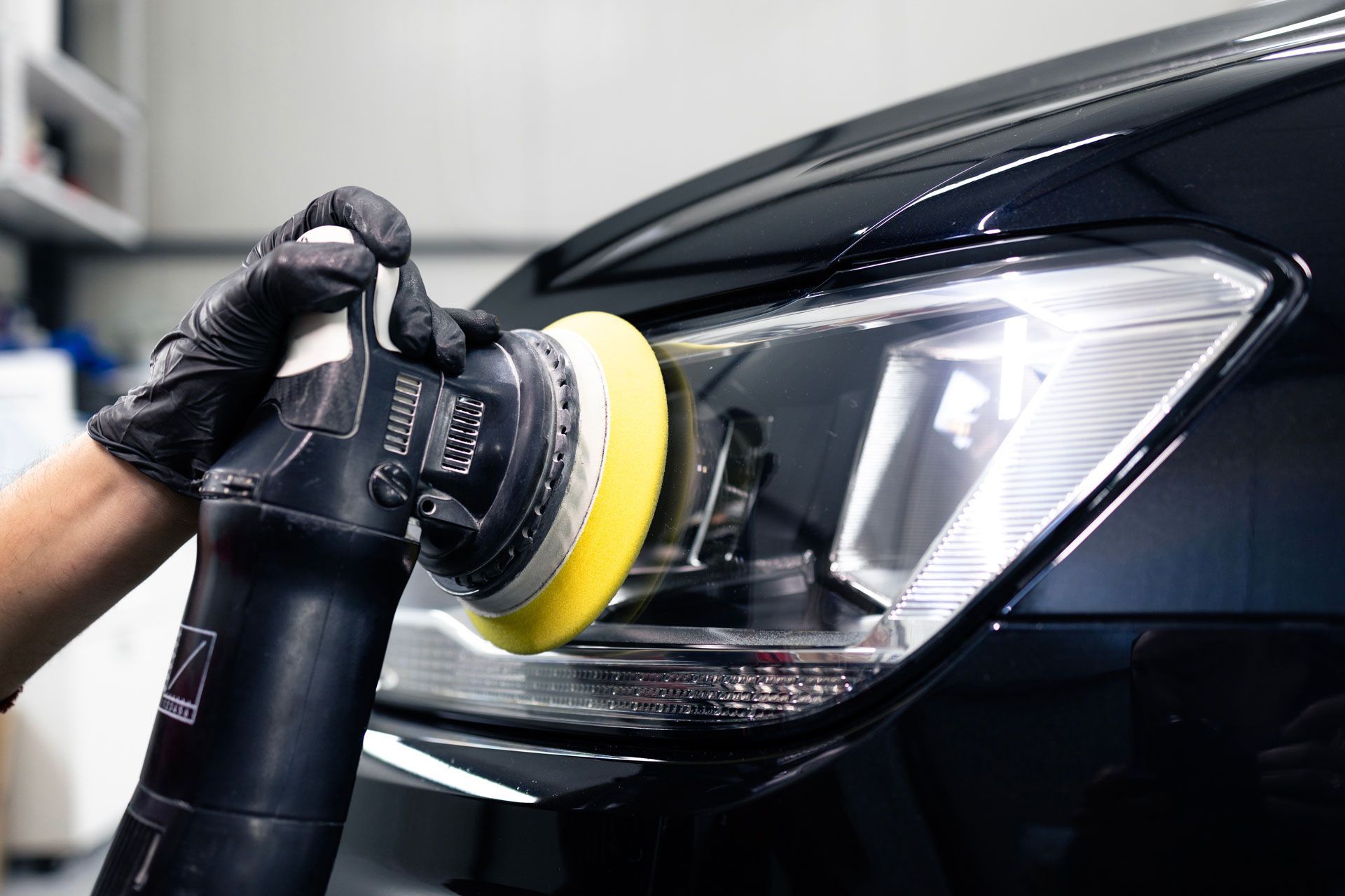 Person polishing a car headlight with a black polishing tool, yellow pad, and black glove.